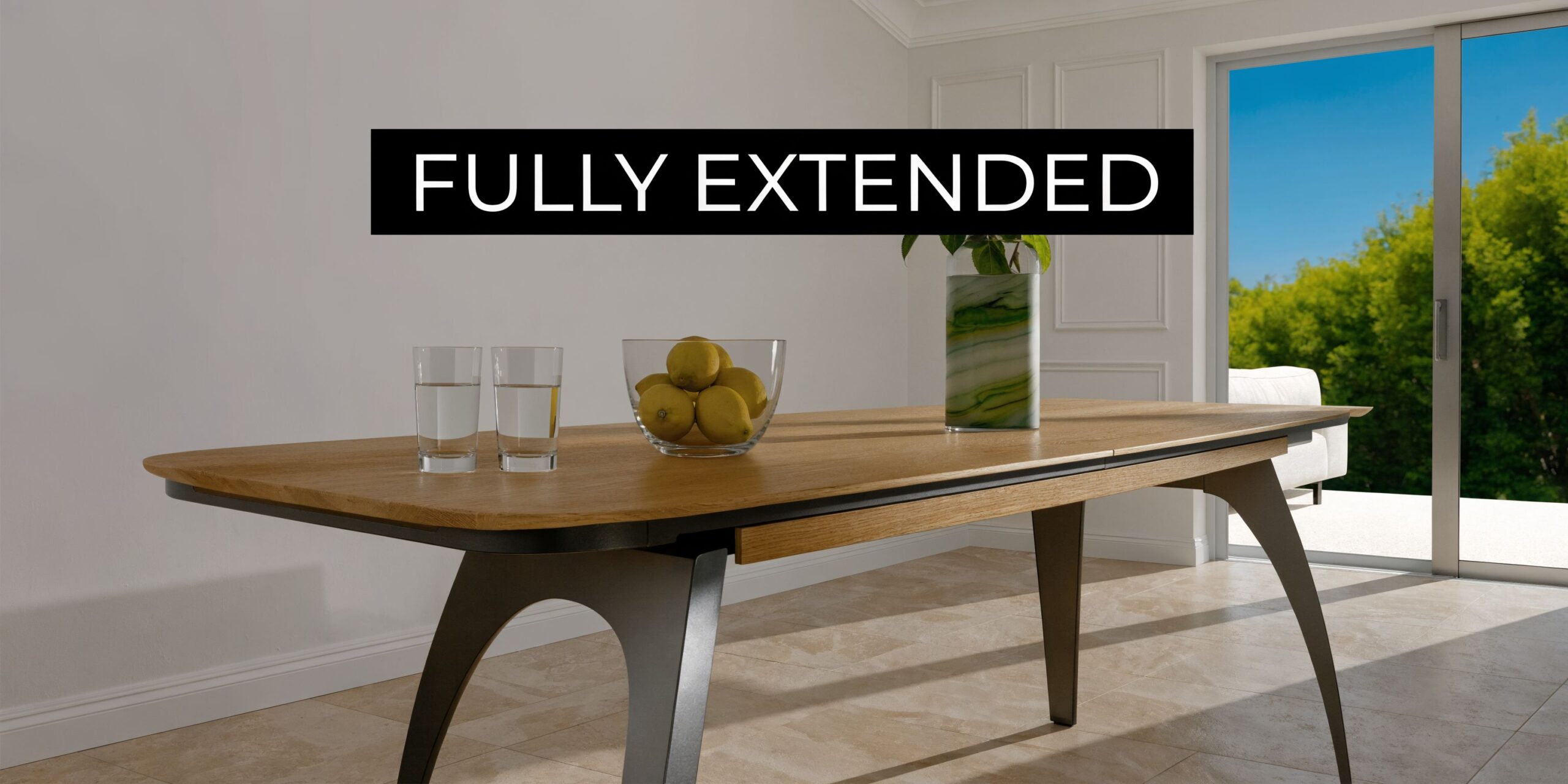 A modern wooden dining table fully extended in a bright sunlit room with glasses and fruit centerpiece.